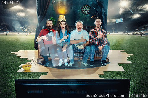 Image of Soccer football fans sitting on the sofa and watching TV in the middle of a football field.