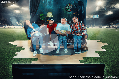 Image of Soccer football fans sitting on the sofa and watching TV in the middle of a football field.