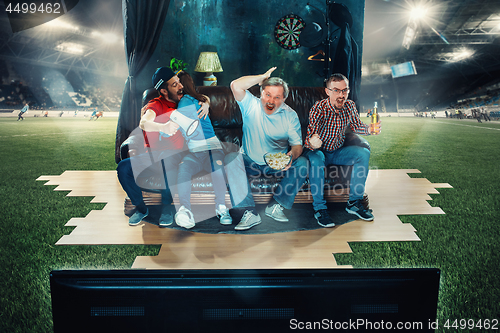 Image of Soccer football fans sitting on the sofa and watching TV in the middle of a football field.