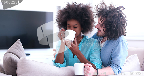 Image of multiethnic couple sitting on sofa at home drinking coffe