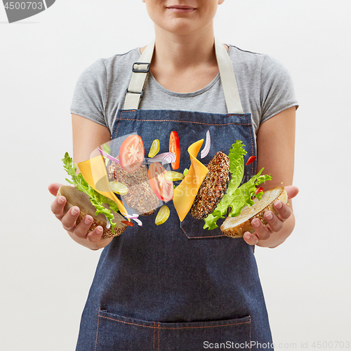 Image of Female chef in a denim apron with flying tasty homemade burger from fresh natural ingredients in her hands on a white background. Place for text.