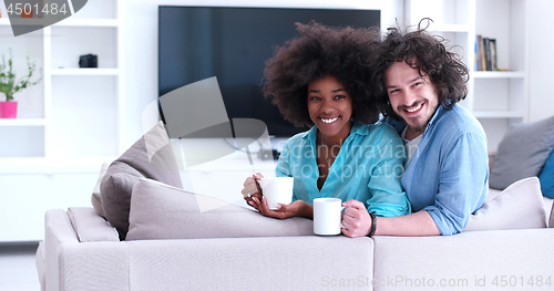 Image of multiethnic couple sitting on sofa at home drinking coffe