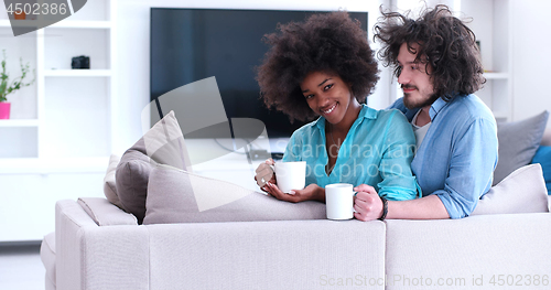 Image of multiethnic couple sitting on sofa at home drinking coffe