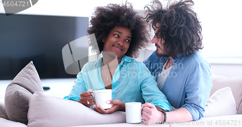 Image of multiethnic couple sitting on sofa at home drinking coffe