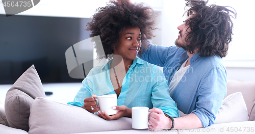 Image of multiethnic couple sitting on sofa at home drinking coffe