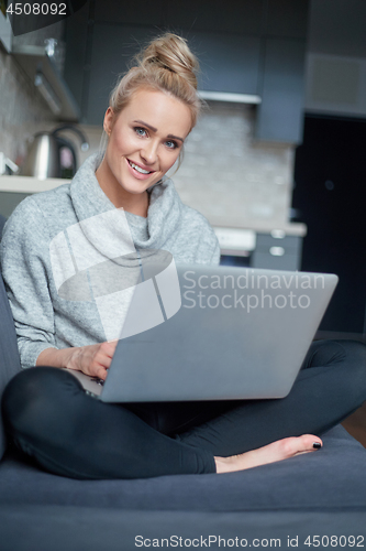 Image of Cheerful young blond woman sitting on couch in living room and using laptop