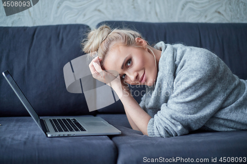 Image of Happy blond woman lying prone on sofa and working on laptop computer