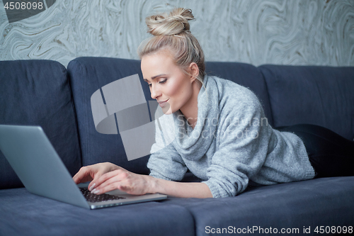 Image of Happy blond woman lying prone on sofa and working on laptop computer
