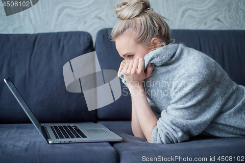 Image of Scaried blond woman lying prone on sofa and looking on laptop computer screen