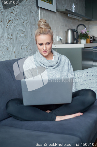 Image of Cheerful young blond woman sitting on couch in living room and using laptop