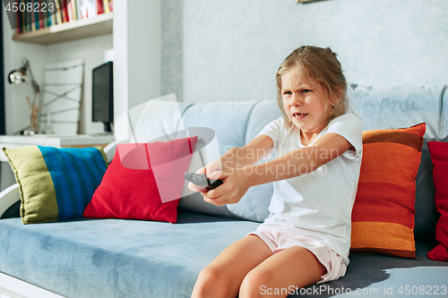 Image of Little casual girl watching tv at home. Female kid sitting on sofa with TV remote and switching channels