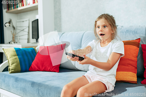 Image of Little casual girl watching tv at home. Female kid sitting on sofa with TV remote and switching channels