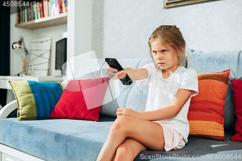 Image of Little casual girl watching tv at home. Female kid sitting on sofa with TV remote and switching channels