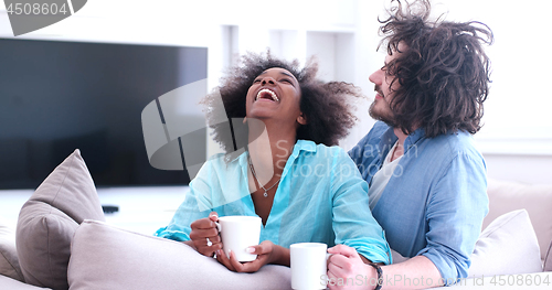 Image of multiethnic couple sitting on sofa at home drinking coffe