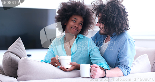 Image of multiethnic couple sitting on sofa at home drinking coffe