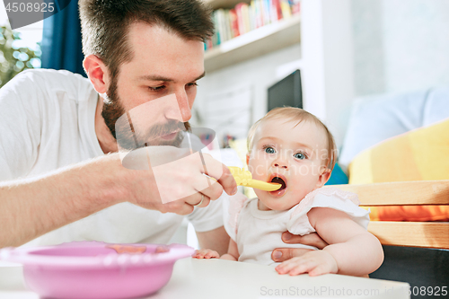 Image of Good looking young man eating breakfast and feeding her baby girl at home