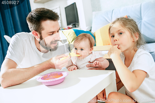 Image of Good looking young man eating breakfast and feeding her baby girl at home