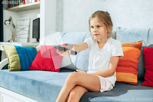 Image of Little casual girl watching tv at home. Female kid sitting on sofa with TV remote and switching channels