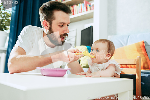 Image of Good looking young man eating breakfast and feeding her baby girl at home