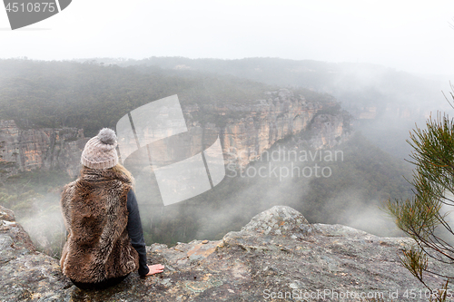 Image of Female sitting on mountain top cliff ledge looking out into the misty fog