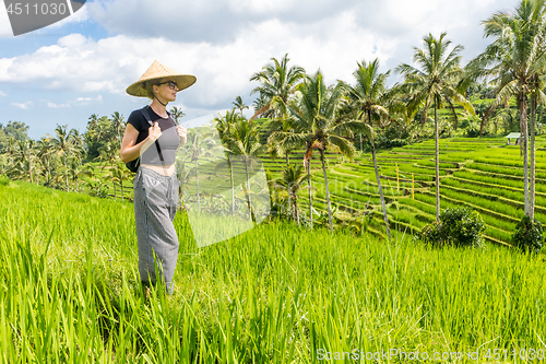 Image of Relaxed fashionable caucasian female tourist wearing small backpack and traditional asian paddy hat walking among beautiful green rice fields and terraces on Bali island