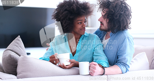 Image of multiethnic couple sitting on sofa at home drinking coffe