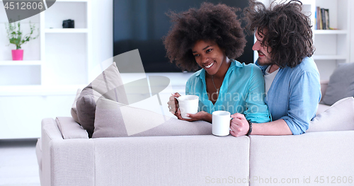 Image of multiethnic couple sitting on sofa at home drinking coffe