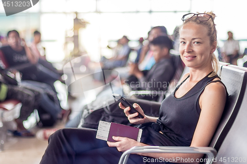 Image of Casual tanned blond female traveler holding cell phone, passport and boarding pass while waiting to board a plane at the departure gates at the asian airport terminal