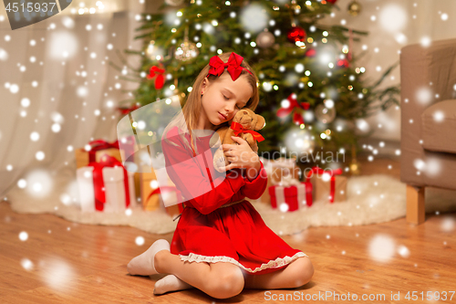 Image of girl in red dress hugging teddy bear at home