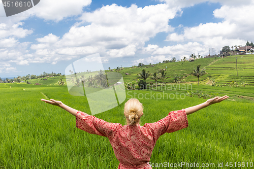 Image of Relaxed fashionable caucasian woman wearing red asian style kimono, arms rised to sky, enjoying pure nature at beautiful green rice fields on Bali island