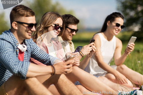 Image of smiling friends with smartphones sitting on grass