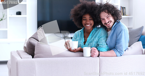 Image of multiethnic couple sitting on sofa at home drinking coffe