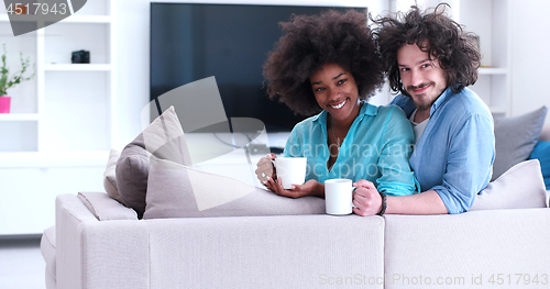 Image of multiethnic couple sitting on sofa at home drinking coffe