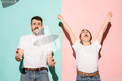 Image of Closeup portrait of young couple, man, woman. One being excited happy smiling, other serious, concerned, unhappy on pink and blue background. Emotion contrasts