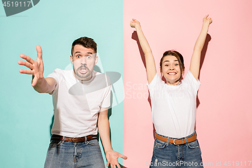 Image of Closeup portrait of young couple, man, woman. One being excited happy smiling, other serious, concerned, unhappy on pink and blue background. Emotion contrasts