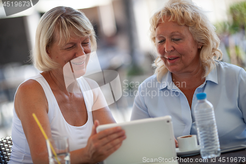 Image of Two smiling senior women at a table of an outdoor cafe looking at the tablet screen