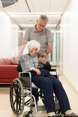 Image of Middle aged man showing and helping elderly 95 years old woman sitting at the wheelchair how to use modern mobile phone.