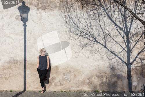 Image of Graphical and textured artisic image of modern trendy fashionable woman wearing sunglasses leaning against old textured retro wall with vintage street lamp and tree shadow falling on the wall.