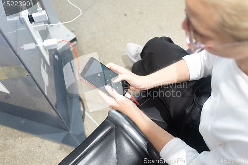 Image of A young woman sitting at a charging station and looking at her smartphone. Recharging mobile phones from free charge station at the airport.