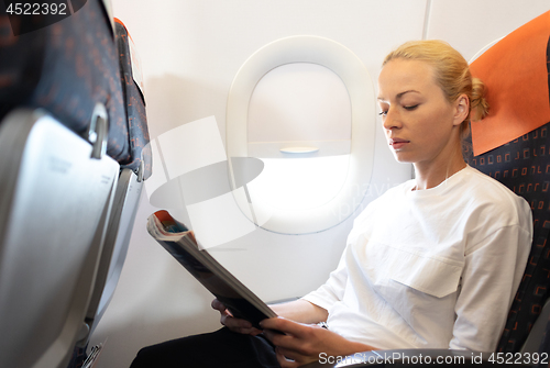 Image of Woman reading in flight magazine on airplane. Female traveler reading seated in passanger cabin. Sun shining trough airplane window.