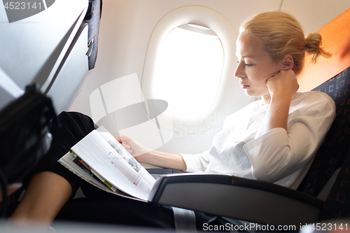 Image of Woman reading in flight magazine on airplane. Female traveler reading seated in passanger cabin. Sun shining trough airplane window