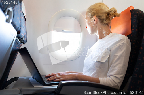 Image of Attractive caucasian female passenger looking through the plain window while working on modern laptop computer using wireless connection on board of commercial airplane flight