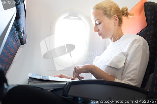 Image of Woman reading in flight magazine on airplane. Female traveler reading seated in passanger cabin. Sun shining trough airplane window