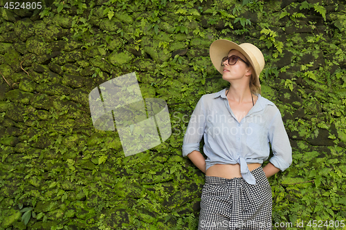 Image of Portrait of a beautiful female traveler. Smiling young woman in summer hat wearing sunglasses, standing in front of lush tropical plant greenery wall background.