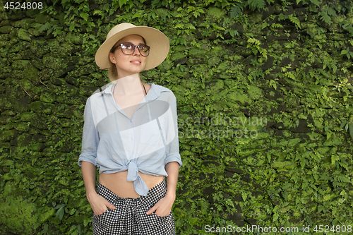 Image of Portrait of a beautiful female traveler. Smiling young woman in summer hat wearing sunglasses, standing in front of lush tropical plant greenery wall background.