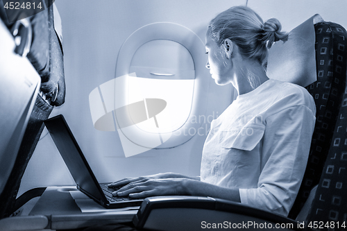 Image of Attractive caucasian female passenger looking through the plain window while working on modern laptop computer using wireless connection on board of commercial airplane flight