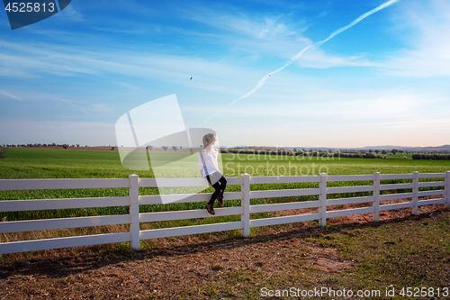 Image of Woman sitting on white farm fence in rural fields