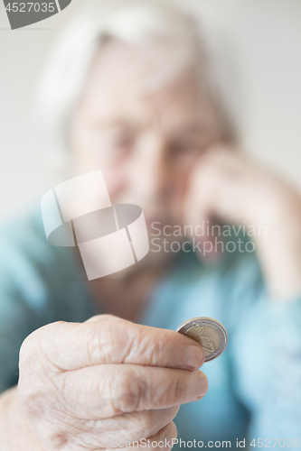 Image of Sad elderly woman sitting at the table at home and looking miserably at only remaining coin from pension in her hand.