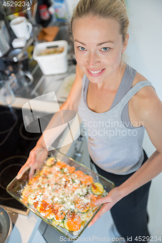 Image of Smiling young healthy woman holding and proudly showing glass baking try with row vegetarian dish ingredients before putting it into oven.