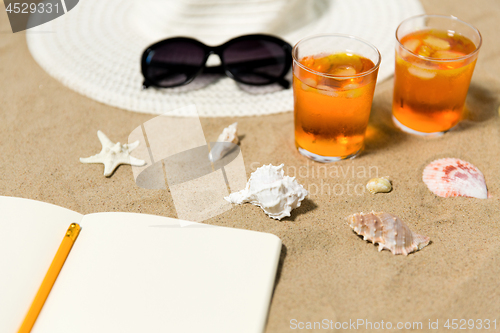 Image of notebook, cocktails, hat and shades on beach sand